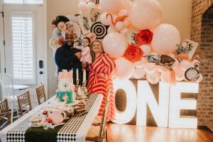 Mother, father, and baby girl in front of our Alice In Wonderland birthday backdrop with custom balloons, red roses, paper fans and decorations over marquee letters, available for your event planned by Just Peachy.